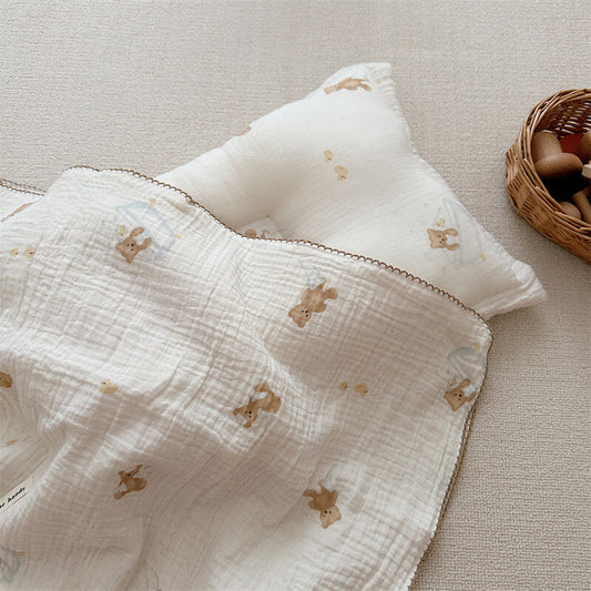 A soft, white cotton muslin toddler blanket with a brown bear print and a matching pillow, resting on a beige carpet.