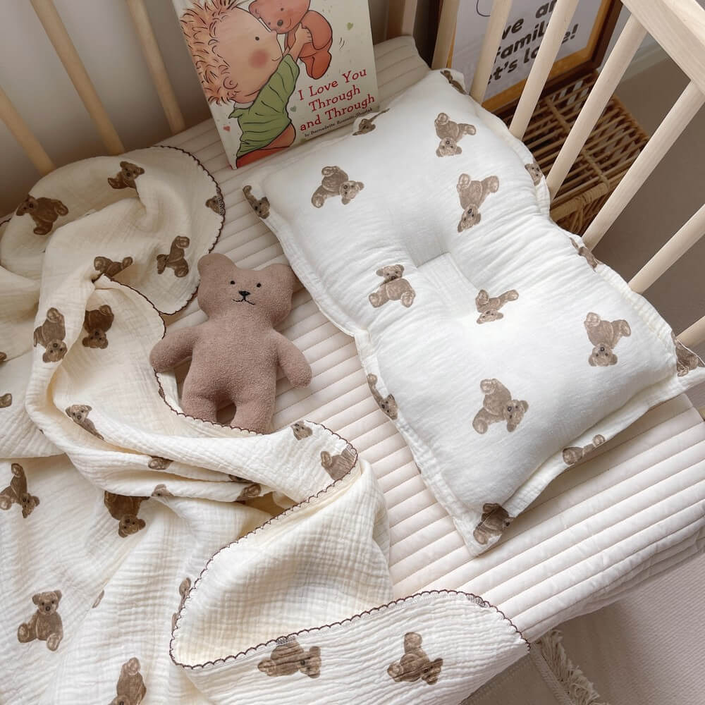 A complete view of the Teddy Bear muslin baby blanket and pillow set arranged in a crib, with a plush toy bear and children's book, showcasing a complete nursery look.