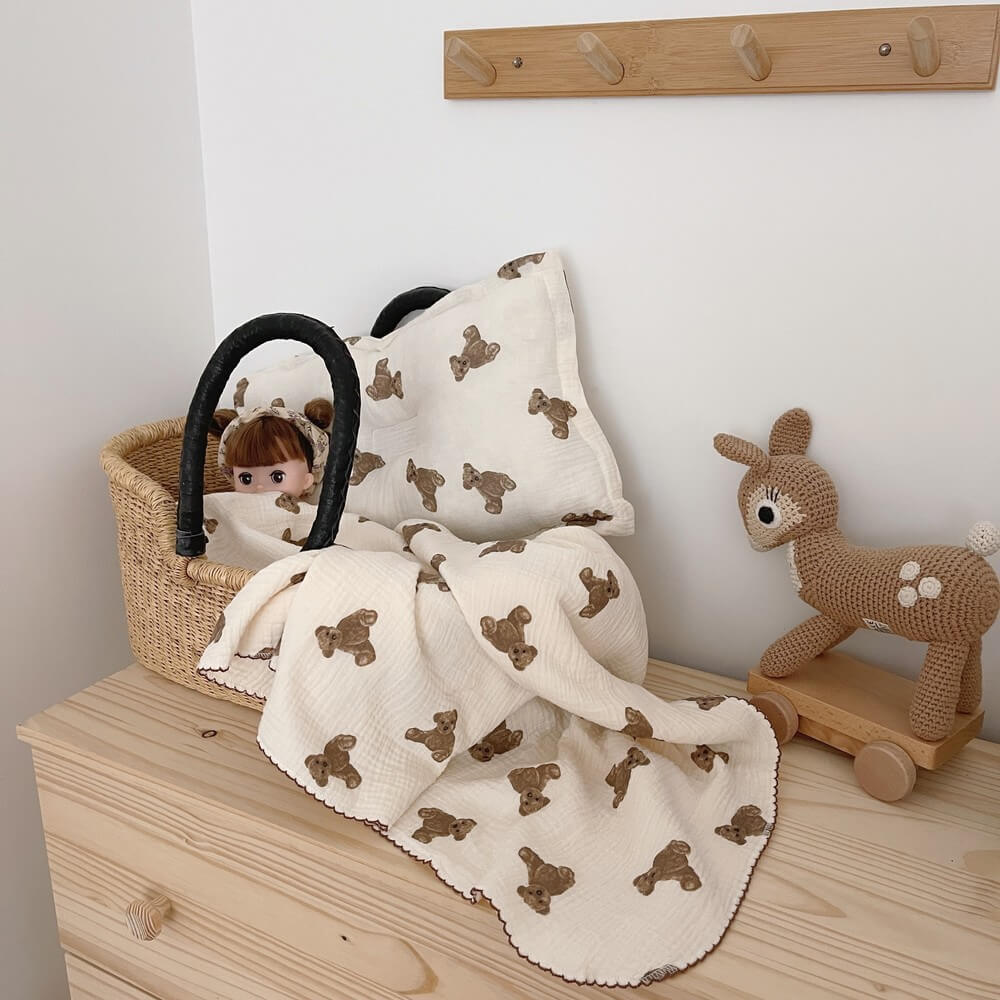 The Teddy Bear muslin baby blanket and pillow set displayed with a doll in a basket, alongside a wooden rocking deer toy. This image shows the full set's charming design.