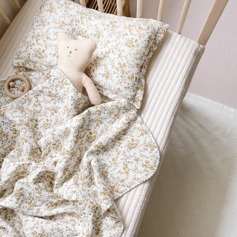 A hibiscus floral cotton muslin blanket and pillow set, suitable for newborns, laid out in a crib with a plush teddy bear and a wooden rattle.