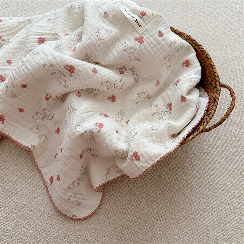 A close-up shot of a white cotton muslin baby blanket with a delicate pattern of gray bunnies and pink flowers