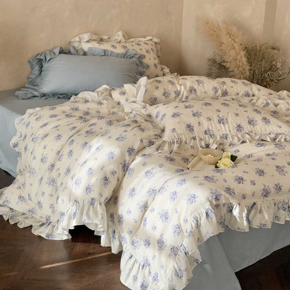 Main product image of the blue cottage rose bedding set on a bed, showcasing the delicate floral pattern, ruffle trim, and the coordinating dusty blue flat sheet.