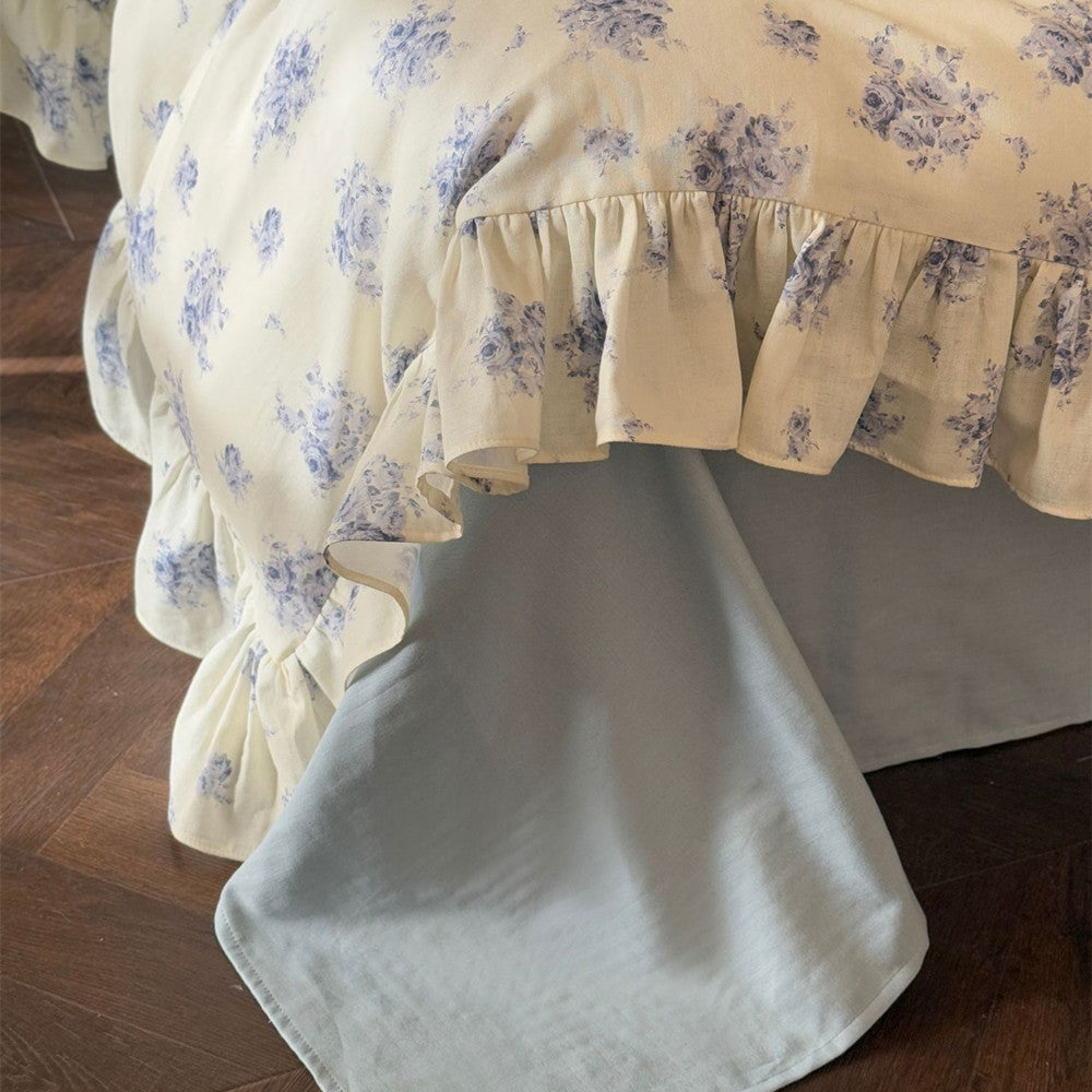 Close-up of the ruffle-trimmed duvet cover, revealing the coordinating solid dusty blue flat sheet peeking out from underneath, showcasing the complete set.