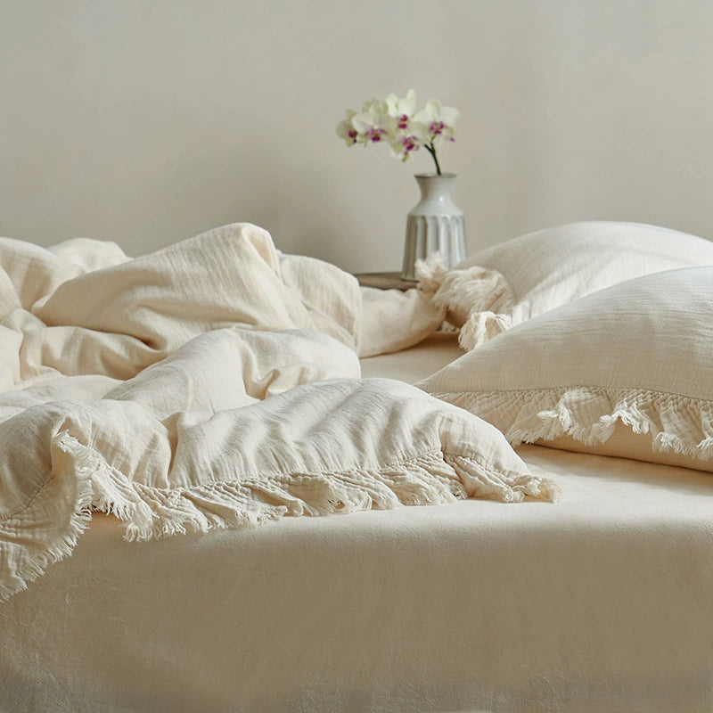 A full-frame view of a cream-colored cotton gauze bedding set, featuring pillows and a duvet with a loose fringe border, captured in soft natural light.