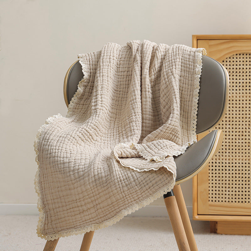 A neutral, light beige 6-layer cotton gauze muslin baby blanket with a decorative lace trim on a gray chair.