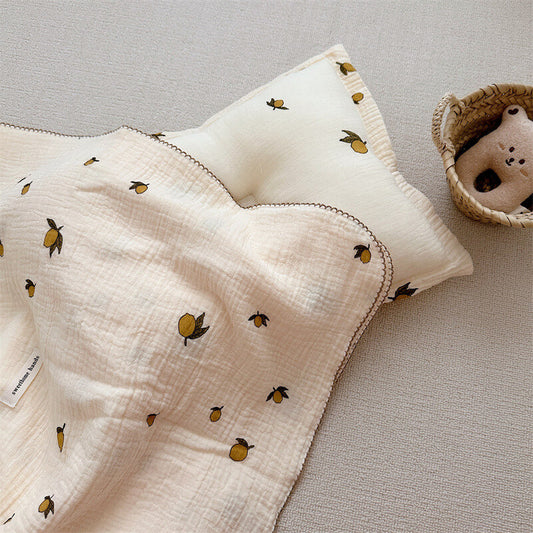 An overhead view of a cotton muslin baby blanket and pillow set, featuring a lemon print, lying on a light-colored carpet with a toy basket in the corner.