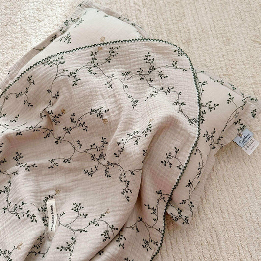 A close-up, top-down view of the Botanical muslin blanket and pillow, highlighting the elegant green vine print and soft, crinkled fabric.