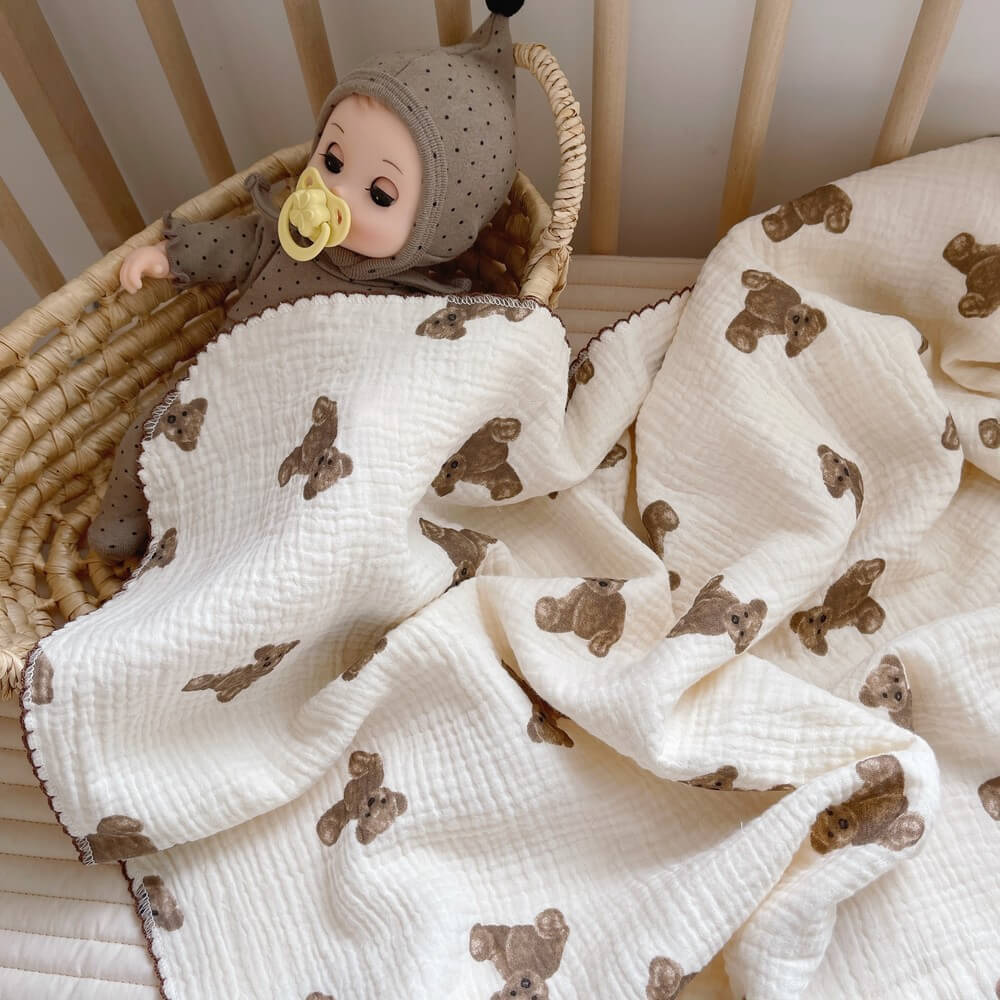 A cute teddy bear muslin baby blanket, 47"x51", draped over a crib with a matching pillow. A baby doll is tucked in, creating a cozy and inviting scene for newborns.