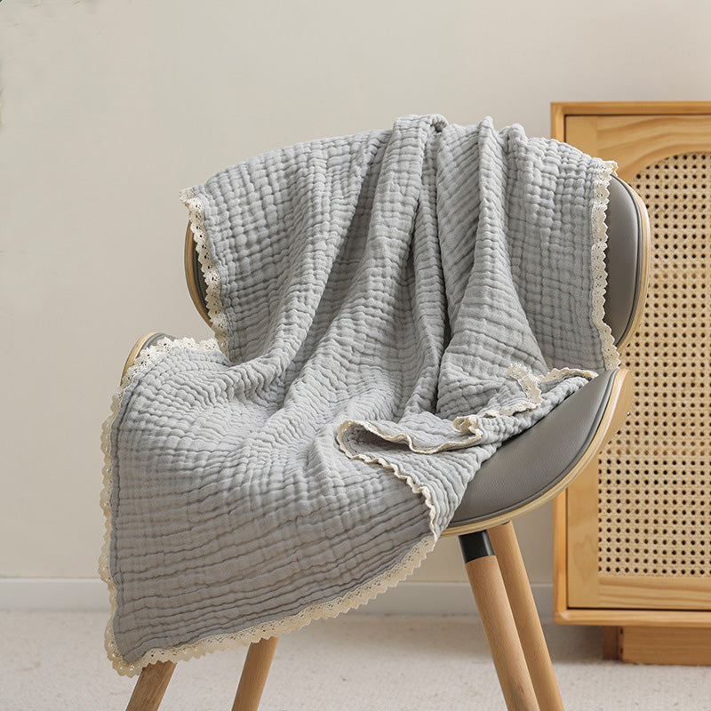 A light gray 6-layer muslin gauze baby blanket with a decorative lace trim draped over a gray chair.