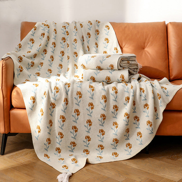 A large ivory acrylic knitted throw blanket with a floral pattern and tassels draped over a leather sofa, with a neatly folded blanket stack next to it