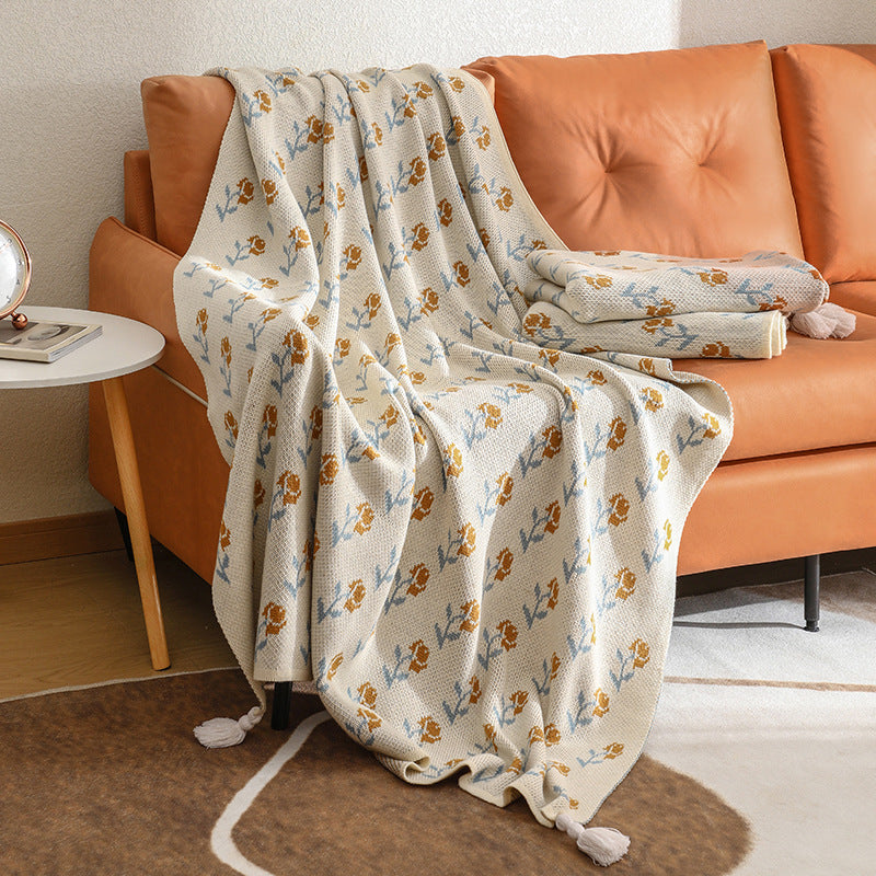 A close-up view of a floral acrylic knitted throw blanket with tassels, highlighting the soft texture and the intricate flower pattern as it drapes over a sofa.