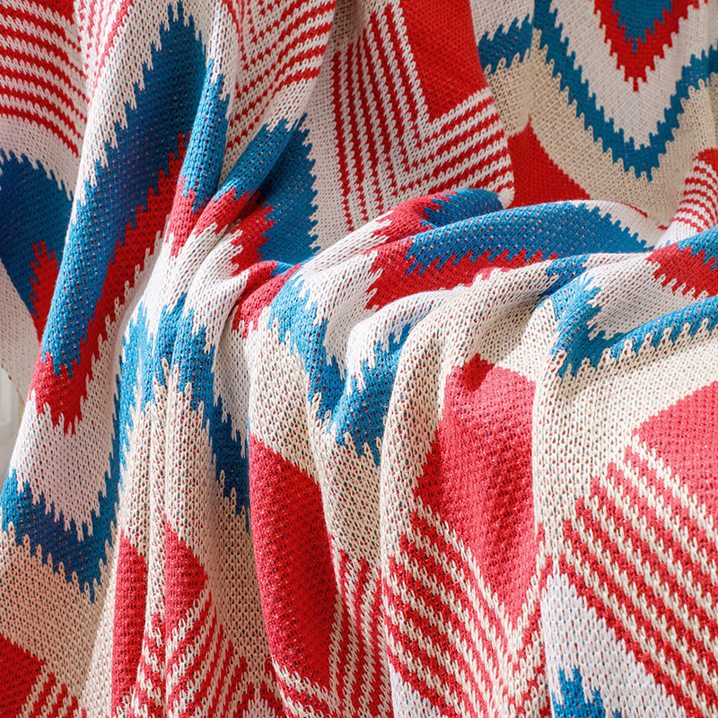 A close-up texture shot of a red, white, and blue chevron patterned knitted throw blanket, showing the detailed weaving and soft folds as it drapes over a sofa.