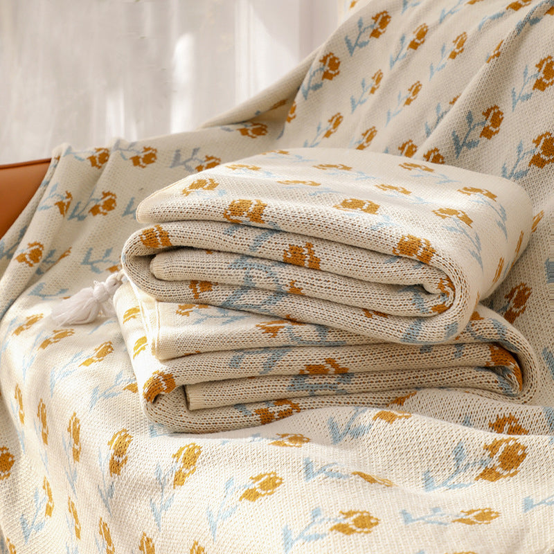 A stack of two ivory floral acrylic knitted throw blankets with tassels, placed on a beige sofa, emphasizing the softness and texture of the material.