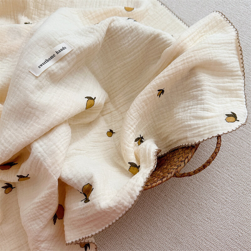 A lightweight, lemon-print muslin baby blanket draped inside a woven basket, showing the textured fabric and delicate hem.