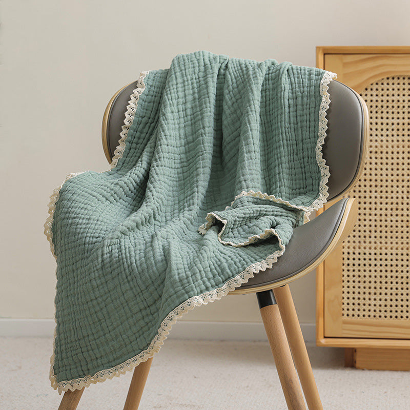 A soft, sage green 6-layer muslin baby blanket with lace trim, perfect for a baby boy, resting on a modern chair.