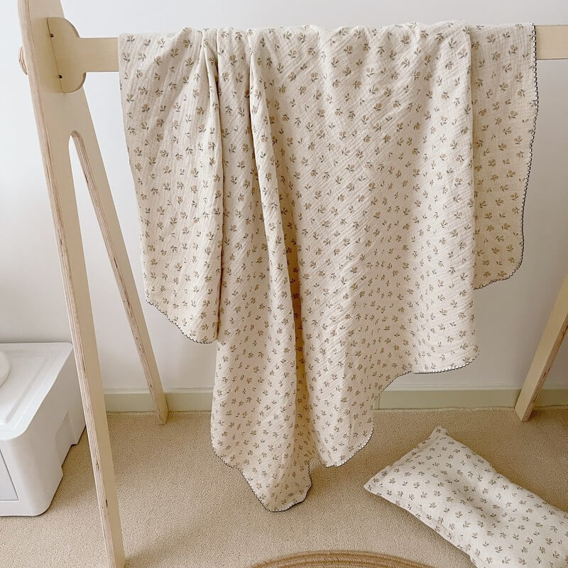 A vintage floral cotton muslin baby blanket and matching pillow draped over a wooden stand, suitable for nursery crib bedding.
