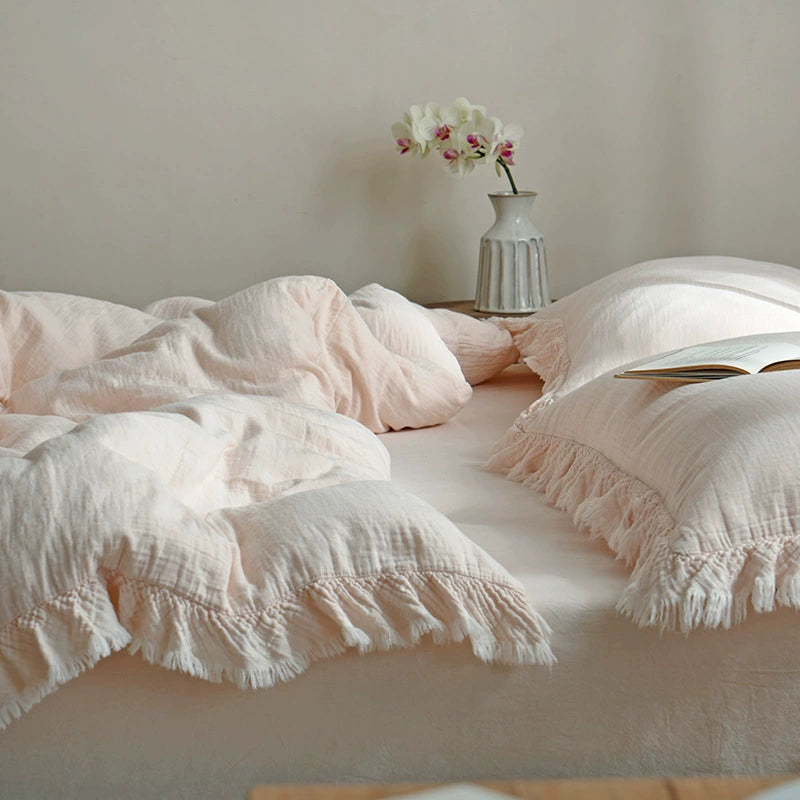 A cozy lifestyle shot of a light pink cotton gauze duvet cover set with fringe and a book resting on the bed, suggesting a relaxing and comfortable space.