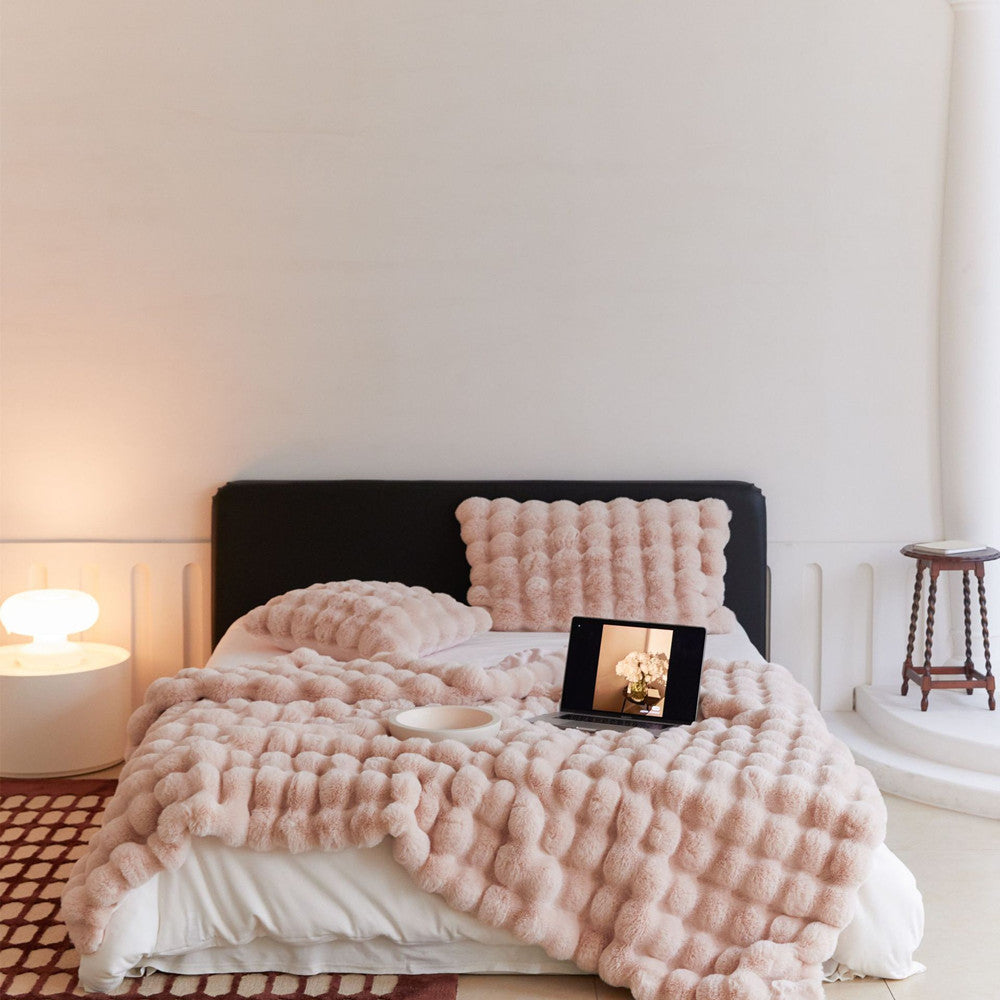 Fluffy blush pink faux rabbit fur blanket and pillows on a bed with a laptop and lamp