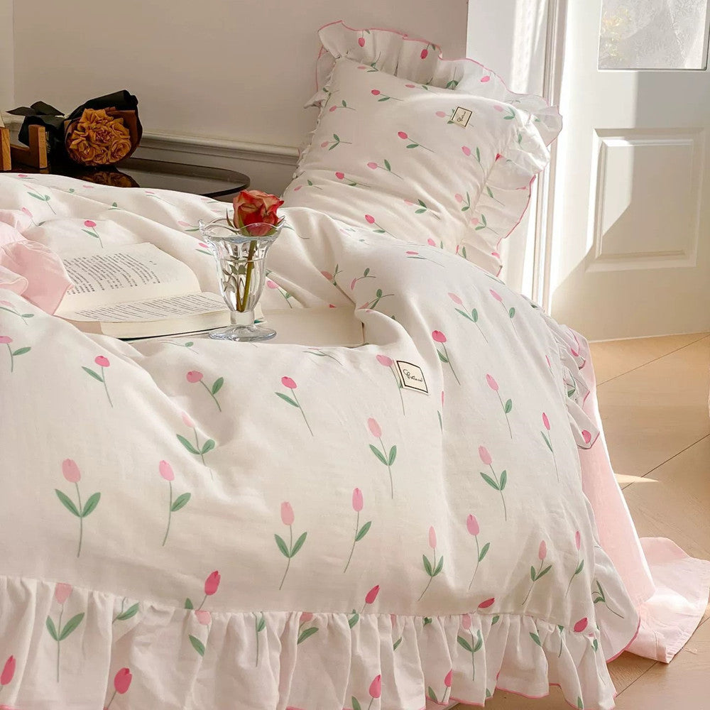 A bed with a pink tulip print bedding set, featuring ruffled trim, styled with an open book and a rose in a vase