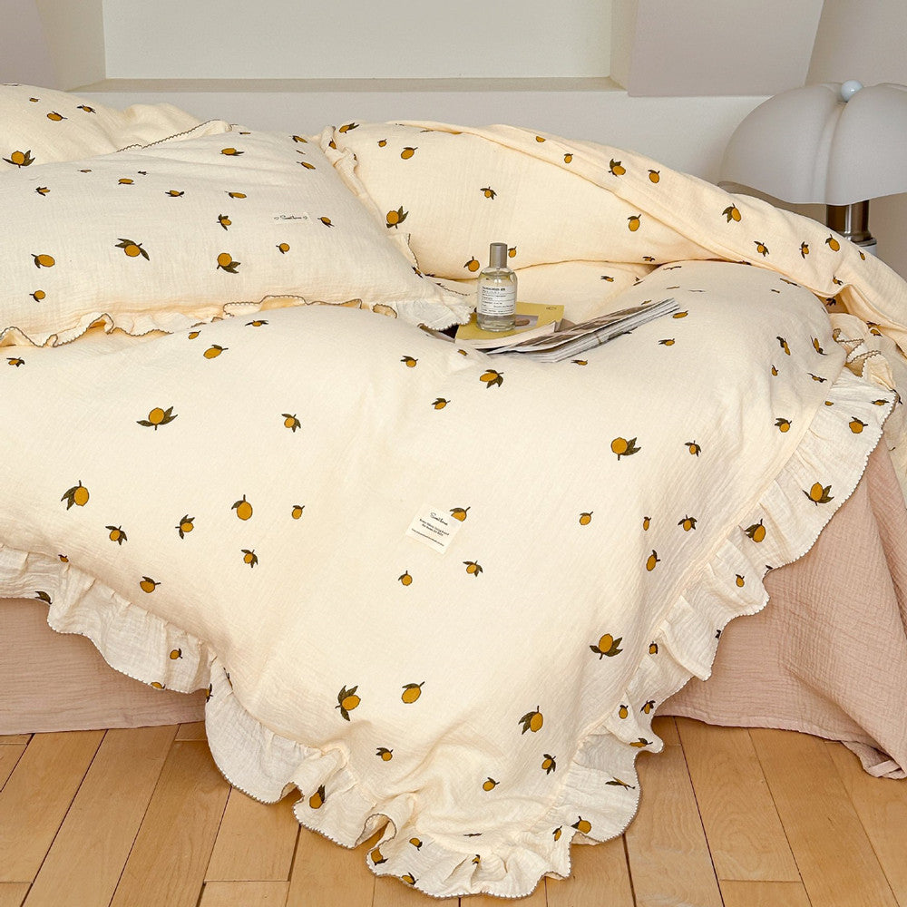 A bed with a Zesty Lemon cotton muslin duvet cover set. The bedding features a cheerful lemon print, ruffled trim, and a small tag with text. A perfume bottle and a magazine are on the bed