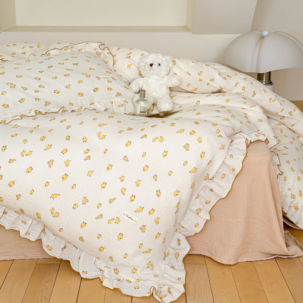A full view of a twin-size bed with a white cotton muslin duvet cover set featuring a delicate duckling print and ruffled trim, a teddy bear rests on the bed