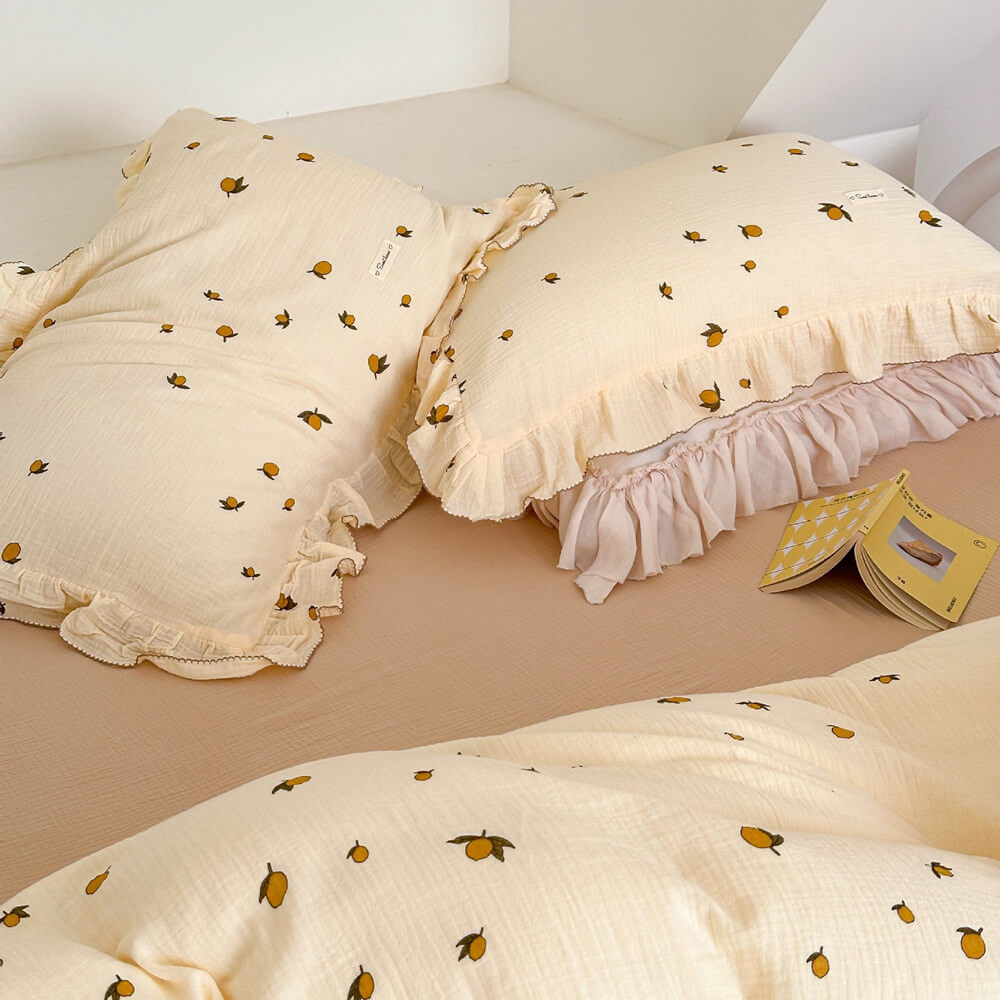 A shot of two pillows from the Zesty Lemon cotton muslin duvet cover set, highlighting the ruffle trim and the delicate lemon print. The pillows are on a tan-colored flat sheet