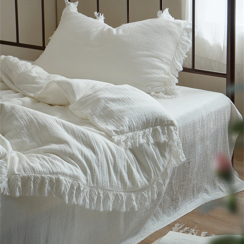 full view of a bed styled with a white cotton gauze duvet cover set, featuring elegant fringe detailing. The bed has a dark metal headboard