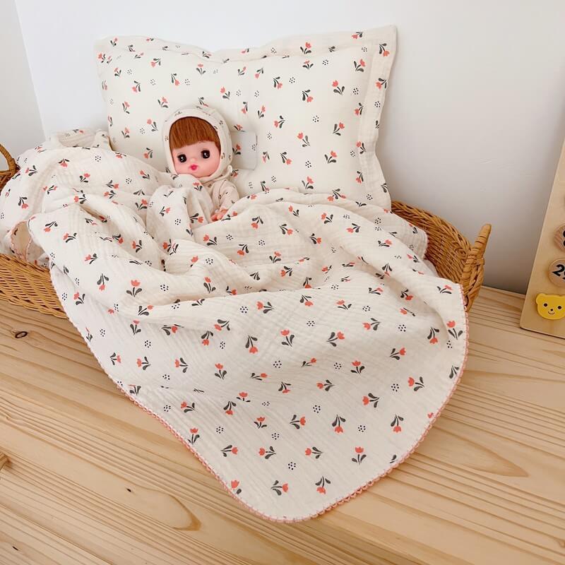 A charming scene with a baby doll nestled in a basket, covered by the soft tulip floral muslin baby blanket and pillow set.