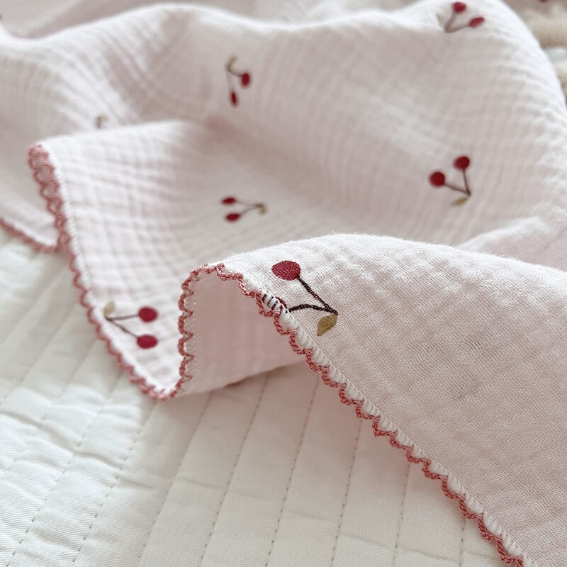 Close-up of a soft cotton muslin baby blanket with an embroidered cherry pattern and scalloped edge.