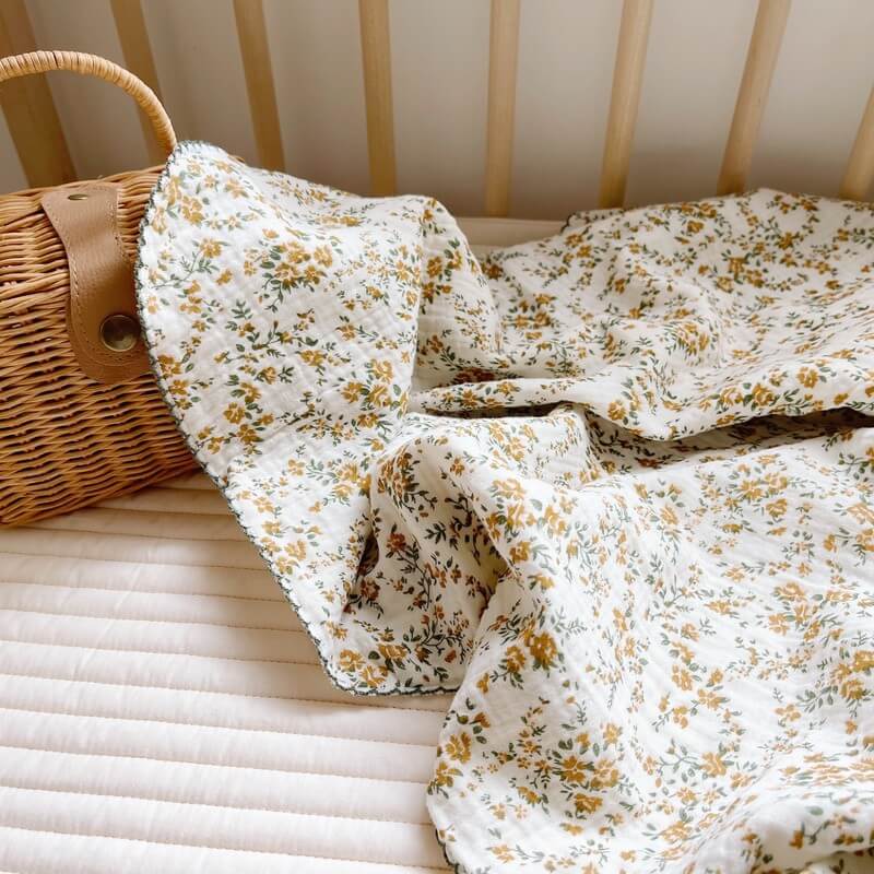 A hibiscus floral cotton muslin swaddle blanket draped over a white quilted crib mattress next to a wicker basket, showing its delicate print