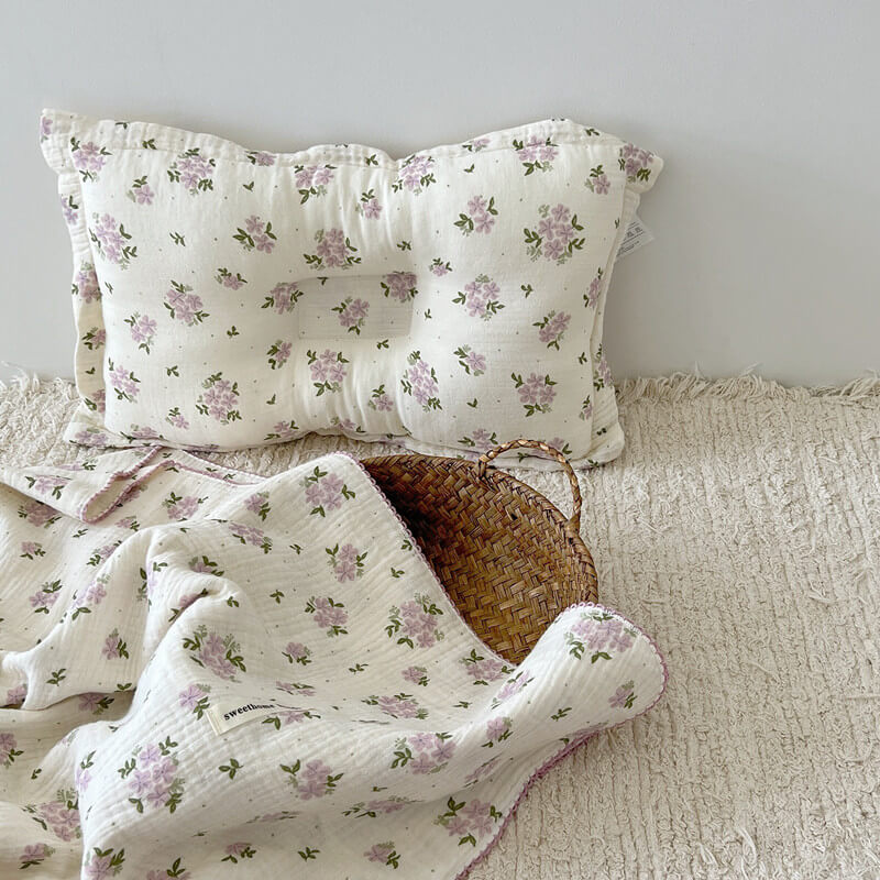 A purple floral reversible cotton gauze baby blanket with a matching pillow, ideal for a crib, on a light-colored woven rug.