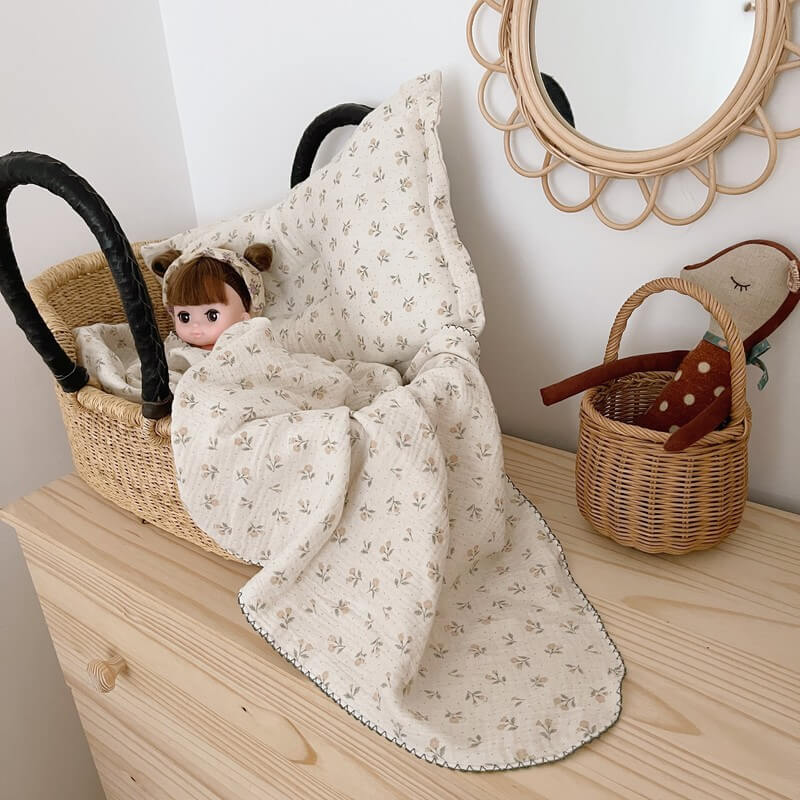 A doll resting in a wicker bassinet, covered with a vintage floral double gauze blanket and pillow, set on a wooden dresser in a nursery