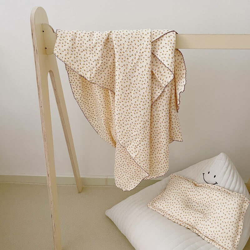 A daisy floral muslin baby blanket and a matching pillow, draped over a wooden stand next to a white bean bag chair.
