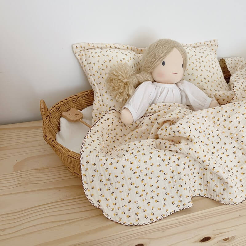 A doll lying in a wicker bassinet, covered with a daisy floral muslin baby blanket and a matching pillow, showing the delicate floral print and scalloped edge.
