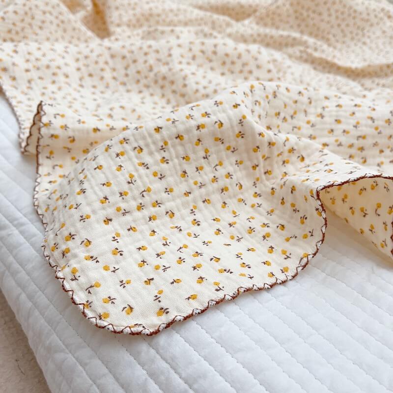 Close-up of a soft, yellow daisy floral muslin baby blanket with a brown scalloped edge, lying on a white quilted surface.