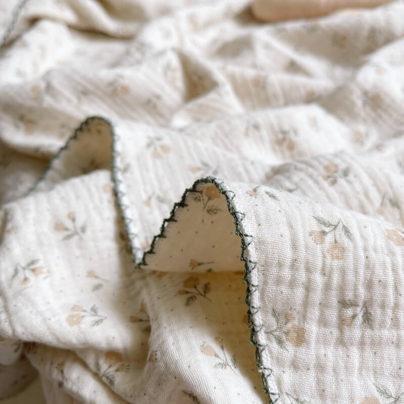 Close-up of a folded gauze swaddle blanket with a vintage floral pattern and a dark green scalloped trim