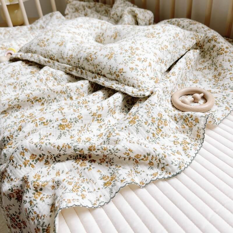 A soft hibiscus floral cotton muslin swaddle blanket and a matching pillow on a white quilted mattress inside a baby crib.