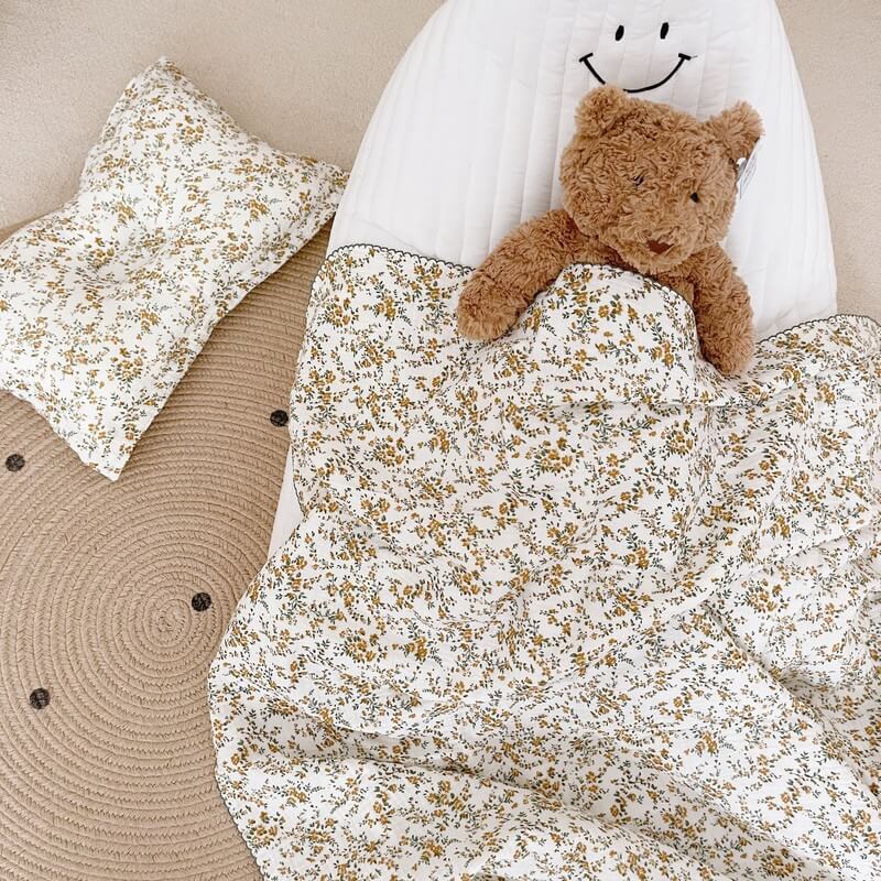 A teddy bear tucked under a hibiscus floral cotton muslin baby blanket, with a matching pillow on a woven rug, a great blanket for infants.