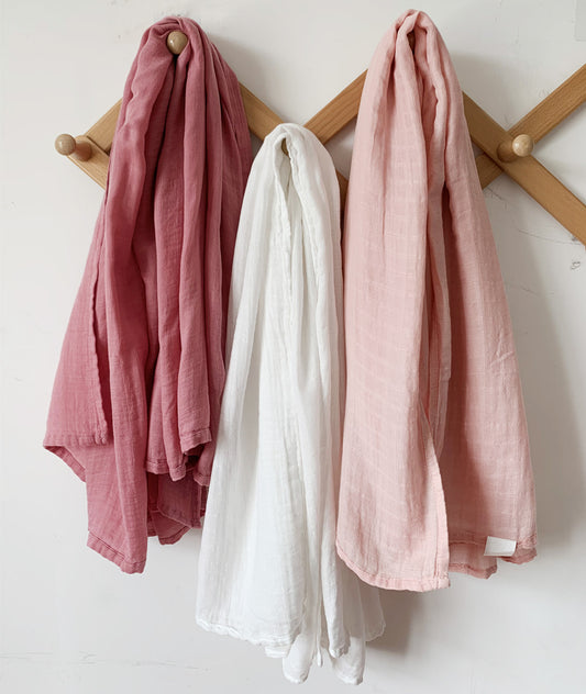 Soft & breathable bamboo muslin swaddle blankets in a 3-pack, featuring dusty rose, off-white, and blush pink colors hanging on a wooden rack