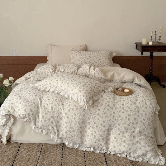 Front view of a full cottagecore bed setup featuring a cream and blue floral print muslin cotton comforter and ruffled lace-trimmed pillows, styled with a rustic bedside table, lit candles, and a vase of white roses.
