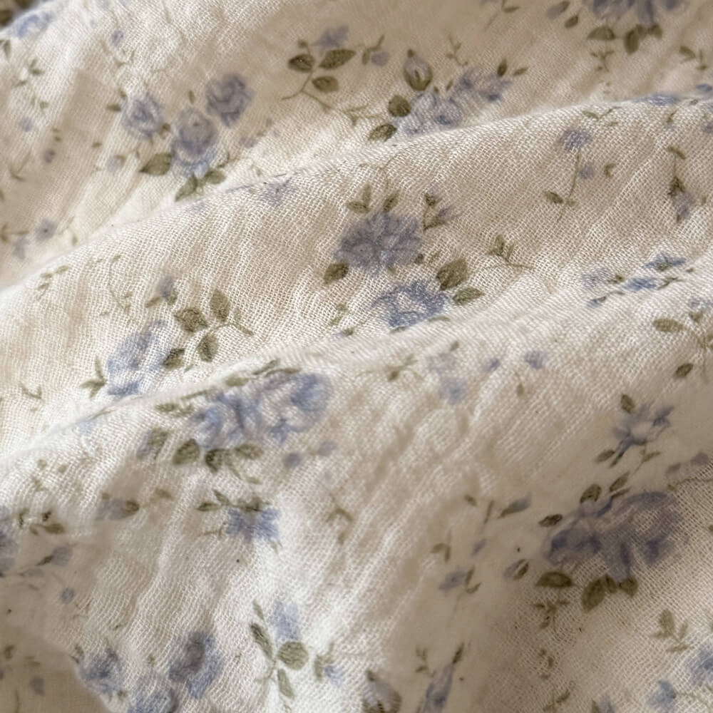 Macro photography showcasing the raw, crinkled texture of unbleached muslin cotton fabric, featuring a high-density delicate blue floral and green leaf print with precise quilting lines.