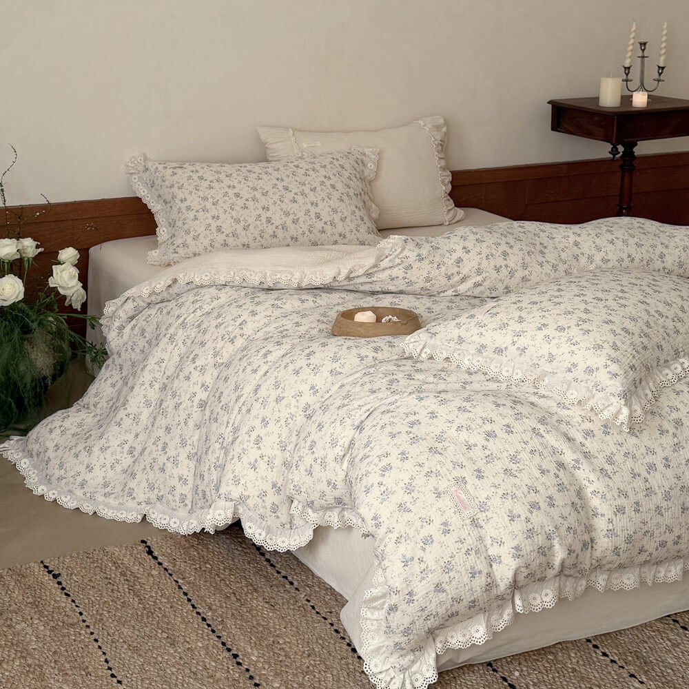 Side angle of a cozy bed dressed with a romantic shabby chic blue floral muslin bedding set, showcasing the comforter's delicate ruffled edges and matching floral pillowcases against a warm wainscot wall.