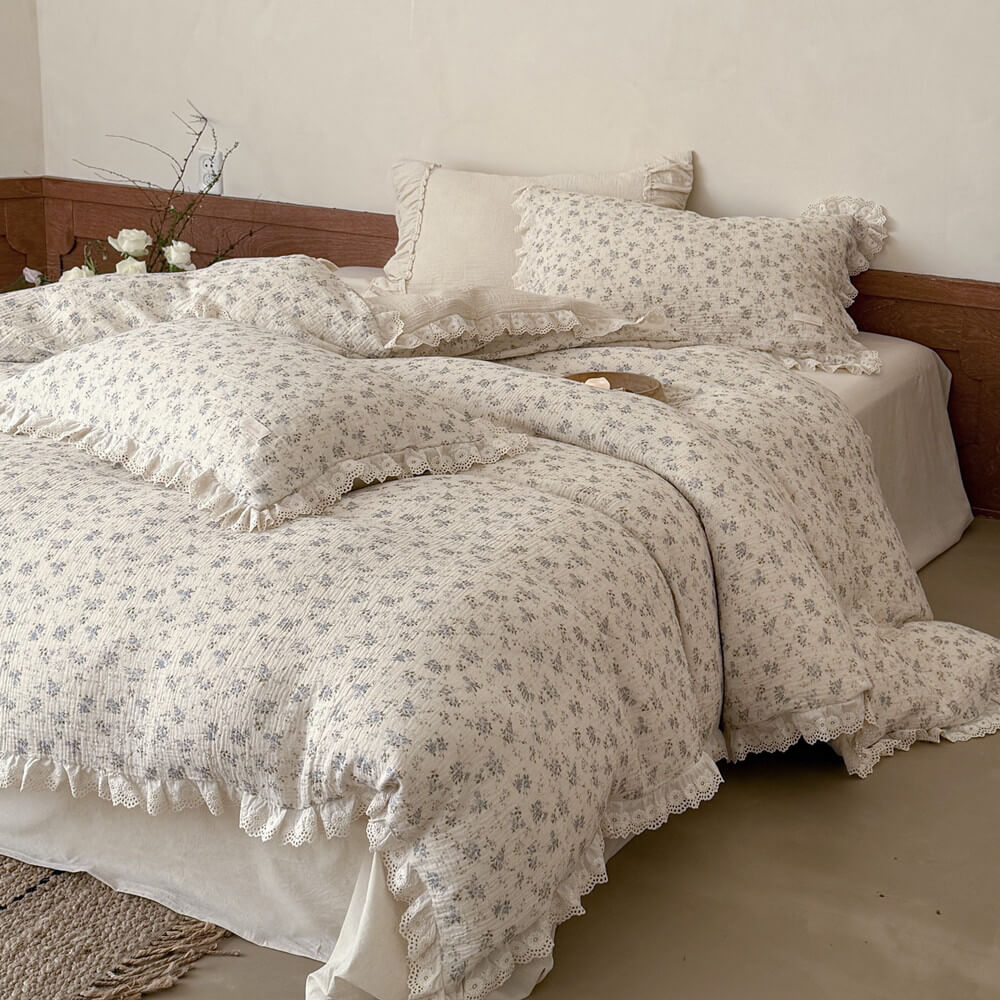 Close-up profile view of a soft, breathable muslin cotton duvet cover in a cream and blue rose pattern, featuring elaborate eyelet lace trim details and natural textile wrinkles.