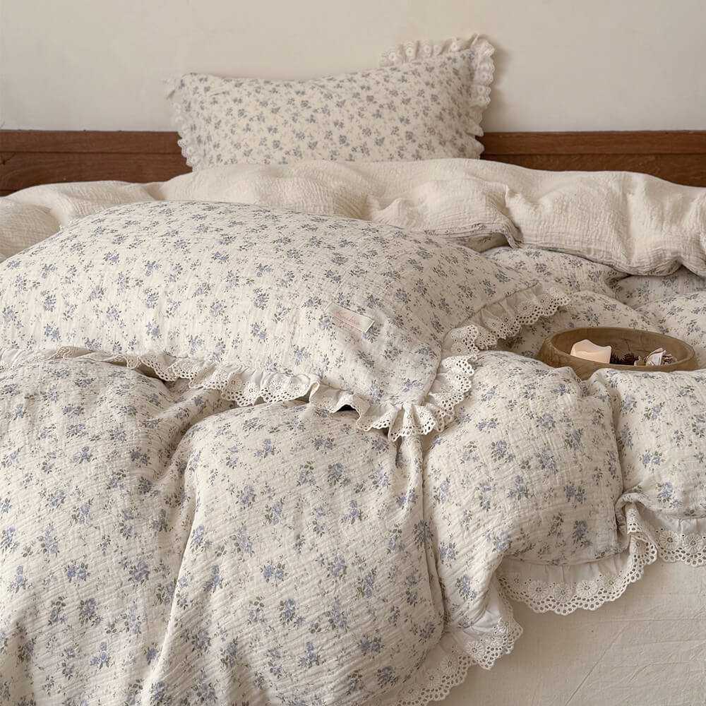 Close-up detail of a wooden tray holding a lit candle resting on a cream muslin quilt with a soft blue floral print and eyelet lace ruffles, emphasizing a relaxing home atmosphere.