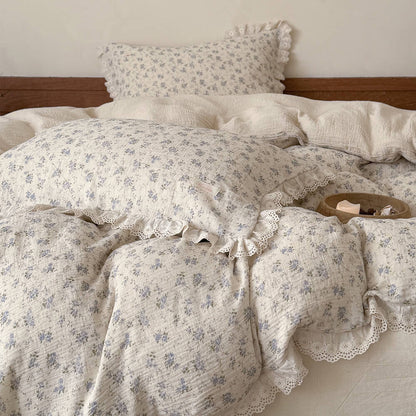 Close-up detail of a wooden tray holding a lit candle resting on a cream muslin quilt with a soft blue floral print and eyelet lace ruffles, emphasizing a relaxing home atmosphere.