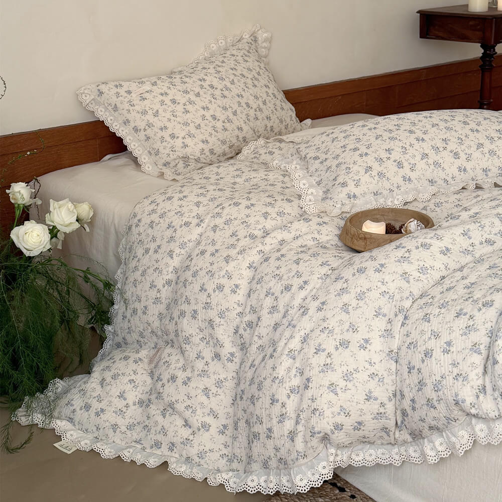 Detailed view of a romantic bedside with a vase of white roses and a lit candle, set against a cottagecore muslin cotton pillow in a matching cream and blue floral print with ruffled edges.