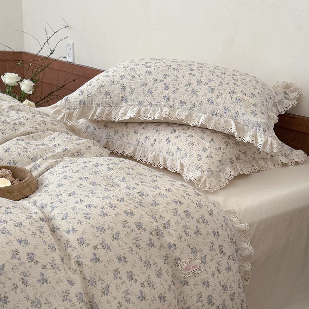 Detail of two stacked shabby chic pillowcases in a crinkled muslin cotton fabric, displaying the unique cream eyelet lace trim and delicate blue rose cottagecore pattern.