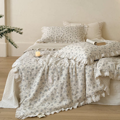 Vintage French style blue floral muslin comforter set draped over a bed, featuring a ruffled eyelet lace trim and matching blue rose cottagecore pillowcases in a cozy, sunlit room.