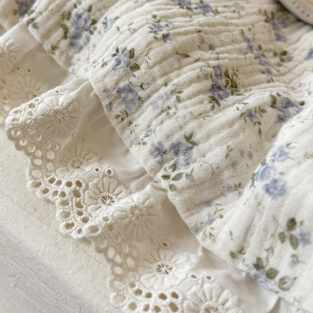 Macro photography of the high-quality embroidered eyelet lace border and the breathable, wrinkled texture of a blue floral muslin cotton duvet cover or comforter fabric.
