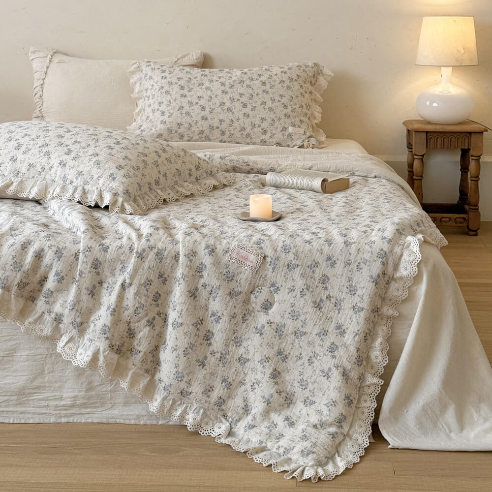 Cottagecore shabby chic bedding set with blue floral printed muslin comforter and ruffled lace pillow shams, styled with a burning candle on a cream-colored bed.